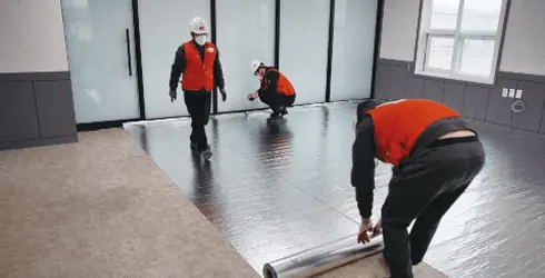 Workers in red vests install insulation sheets using the method B panel in a room with large glass doors and windows, preparing for the Hoya Ondol underfloor heating system. - Hoya Ondol Aust