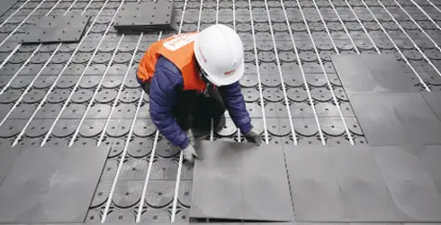 A worker wearing a helmet and vest installs tiles on a grid-like surface, utilizing the install method B panel technique. - Hoya Ondol Aust