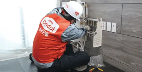 A technician in a red vest and white helmet works on a wall-mounted electric panel in a modern room, ensuring the install method B panel is correctly integrated with the Hoya Ondol underfloor heating system. - Hoya Ondol Aust