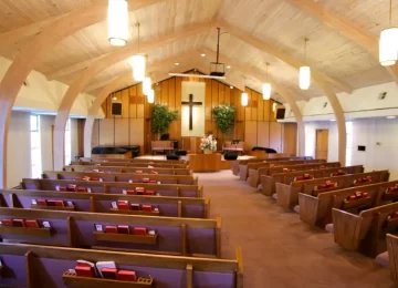 An empty church interior with wooden pews, a large cross at the altar, and soft lighting from hanging fixtures is now comfortably warm, thanks to discreet Hoya Ondol Heating Panels. - Hoya Ondol Aust