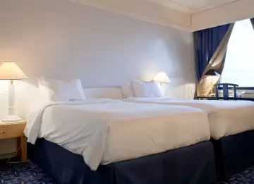 Hotel room with two large beds, white linens, two bedside lamps, and a blue curtain partially open to a window. The cozy ambiance is enhanced by the efficient Hoya Ondol Heating Panels, ensuring warmth and comfort year-round. - Hoya Ondol Aust